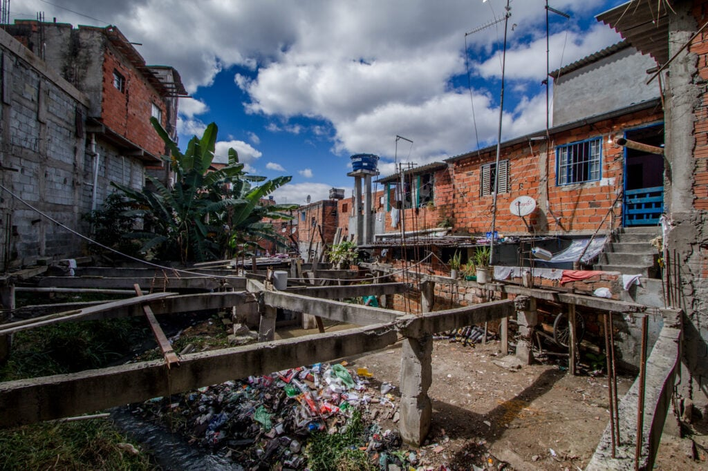 Vala de esgoto a céu aberto em rua de periferia urbana brasileira, com moradias ao fundo