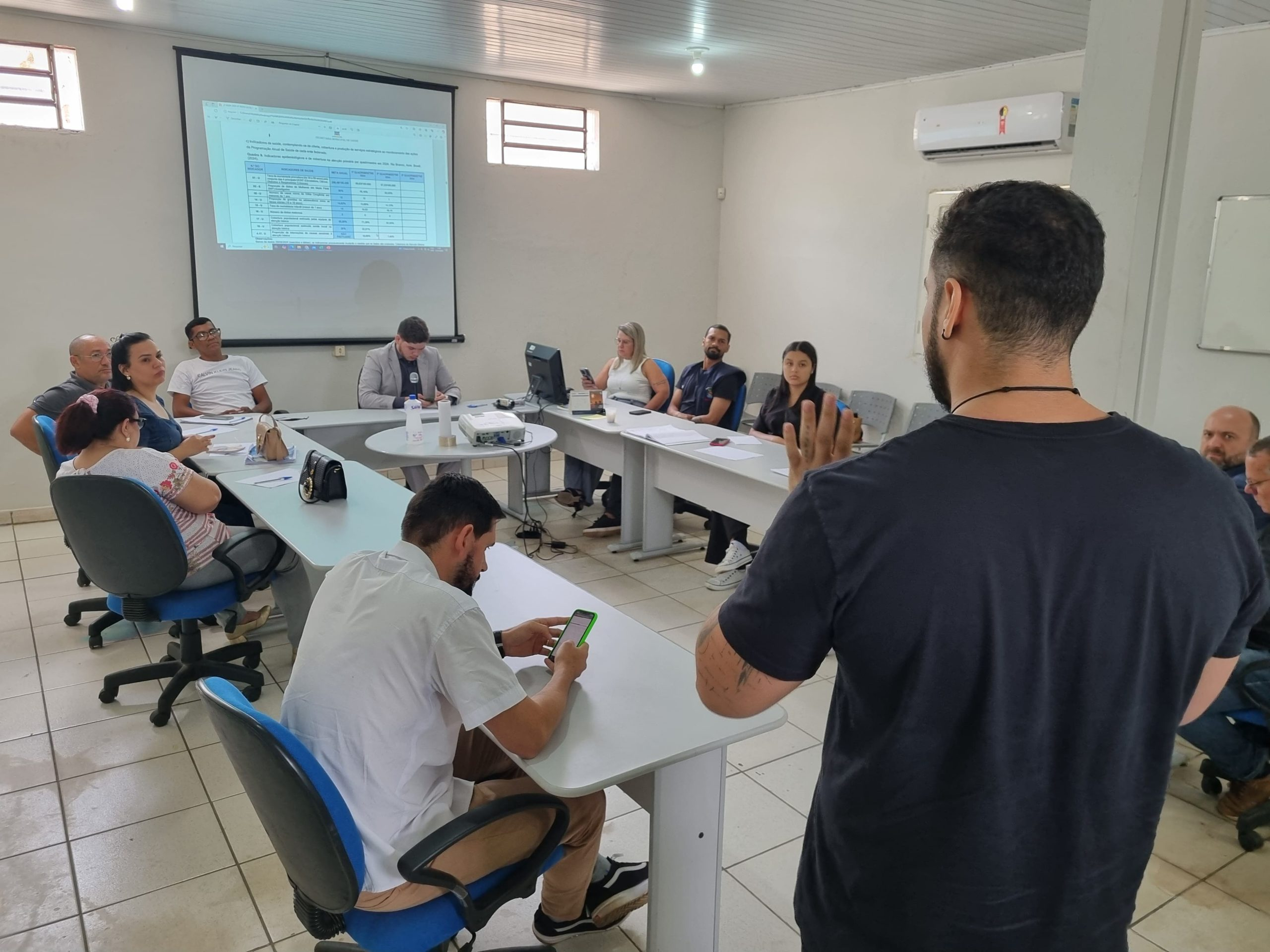Reunião de conselho municipal de saúde com conselheiros sentados em mesa e público presente
